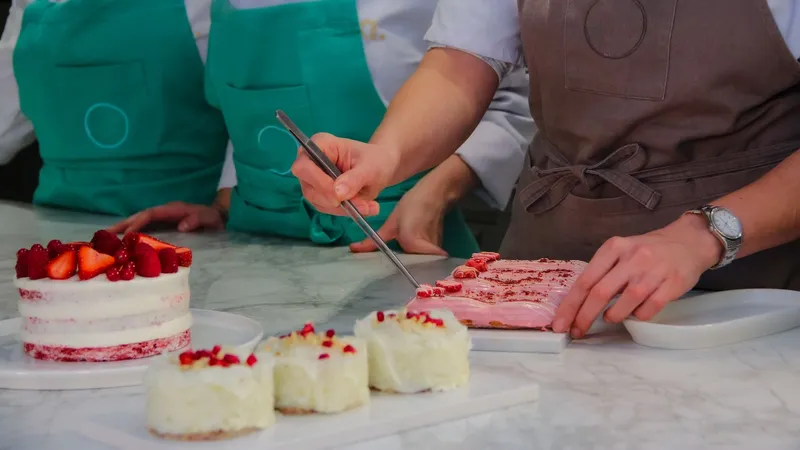 Trois pâtissiers en tablier autour d’un plan de travail en marbre, l’un dressant un entremets rose à la pince devant des gâteaux aux fruits rouges