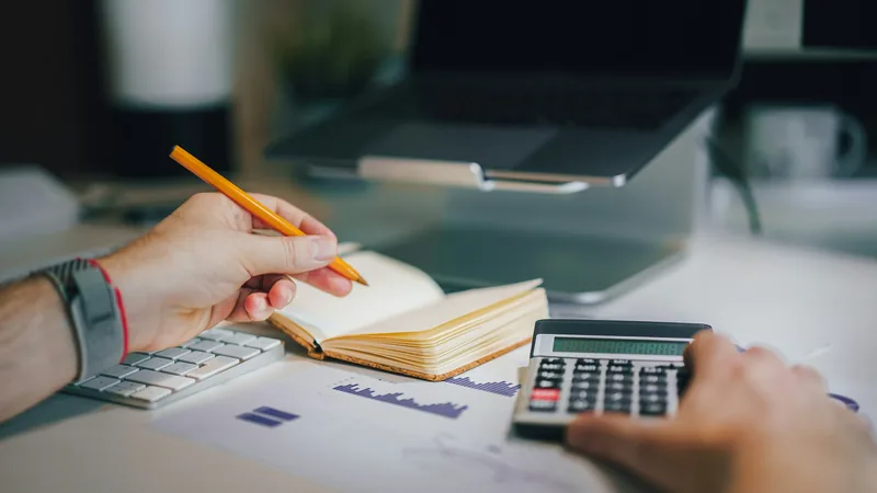 Mains tenant un crayon au-dessus d’un carnet ouvert, à côté d’une calculatrice et d’un graphique imprimé