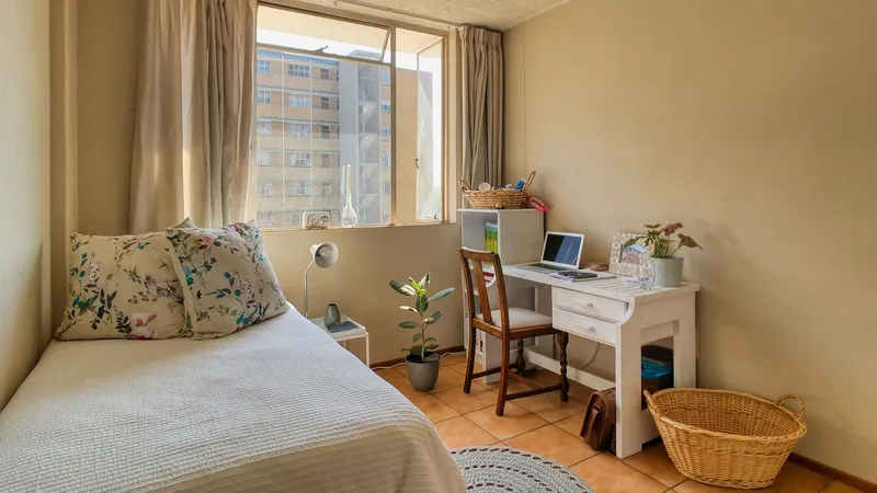 Une chambre d’étudiant lumineuse avec un lit simple, un bureau blanc, un ordinateur portable et une plante près de la fenêtre