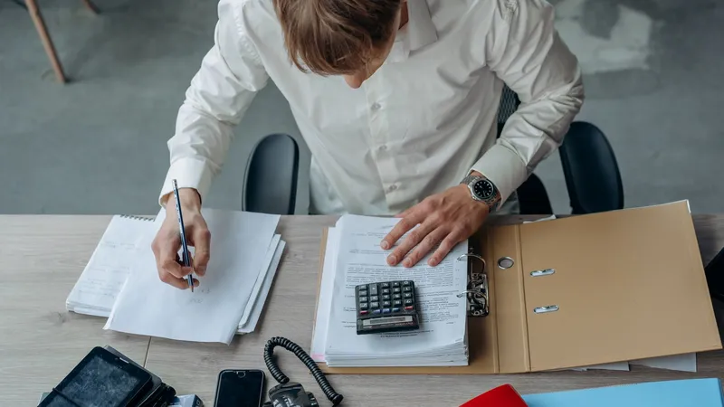 Vu du dessus, un homme en chemise blanche annote des documents à son bureau, devant un classeur ouvert et une calculatrice