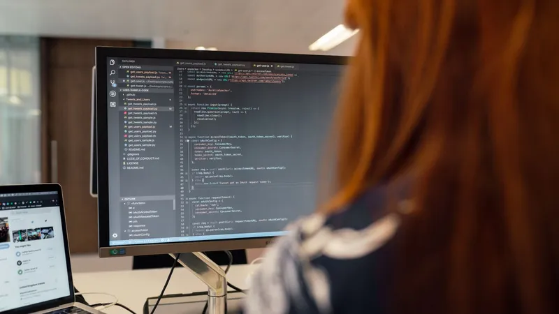 Une développeuse de dos relit du code JavaScript affiché sur un grand écran, à côté d’un ordinateur portable