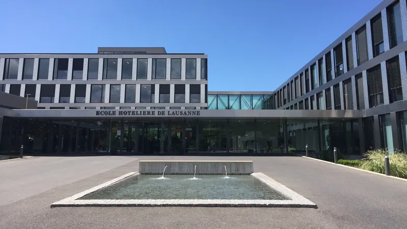 Entrée de l’École hôtelière de Lausanne, bâtiment vitré aux façades claires devant un bassin et ses jets d’eau, sous un ciel bleu