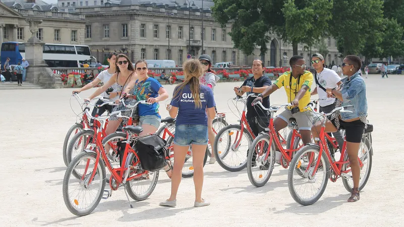 Une guide en tee-shirt bleu s’adresse à un groupe de touristes à vélo sur une esplanade parisienne, devant des bâtiments en pierre