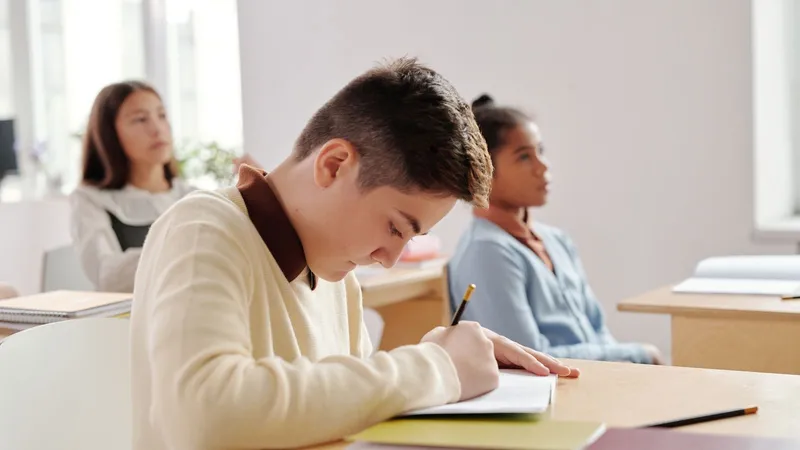 Un lycéen écrit dans son cahier en salle de classe, deux élèves assises derrière lui