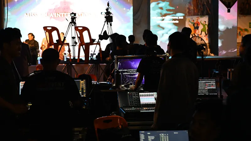 Dans la pénombre, des techniciens règlent les consoles son et vidéo d’un événement, devant une scène éclairée et deux caméras sur trépied